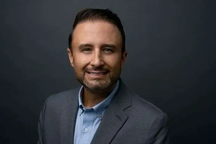 Professional headshot of John Darch, a Birchway Niagara board member, wearing a grey blazer and light blue shirt, smiling against a dark background.