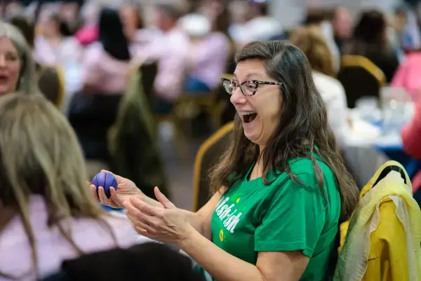 Woman in a green shirt has a big smile sitting at a table while holding a ball.