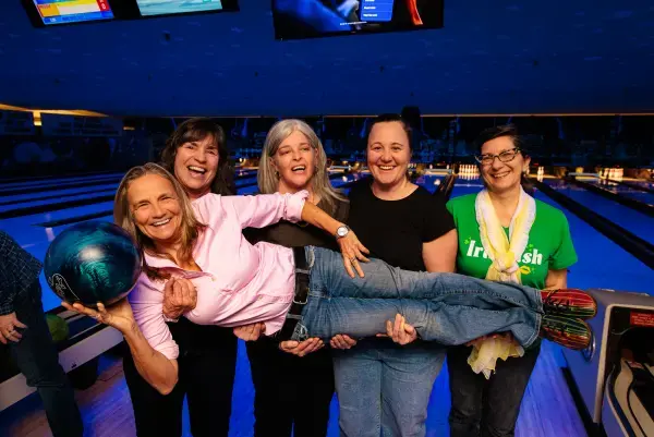 Four women hold a teammate holding a bowling ball in front of their lane