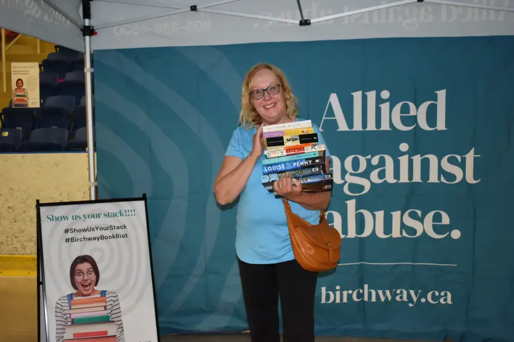 A happy woman holds a stack of books. Behind her is a sign that says Allied against abuse.
