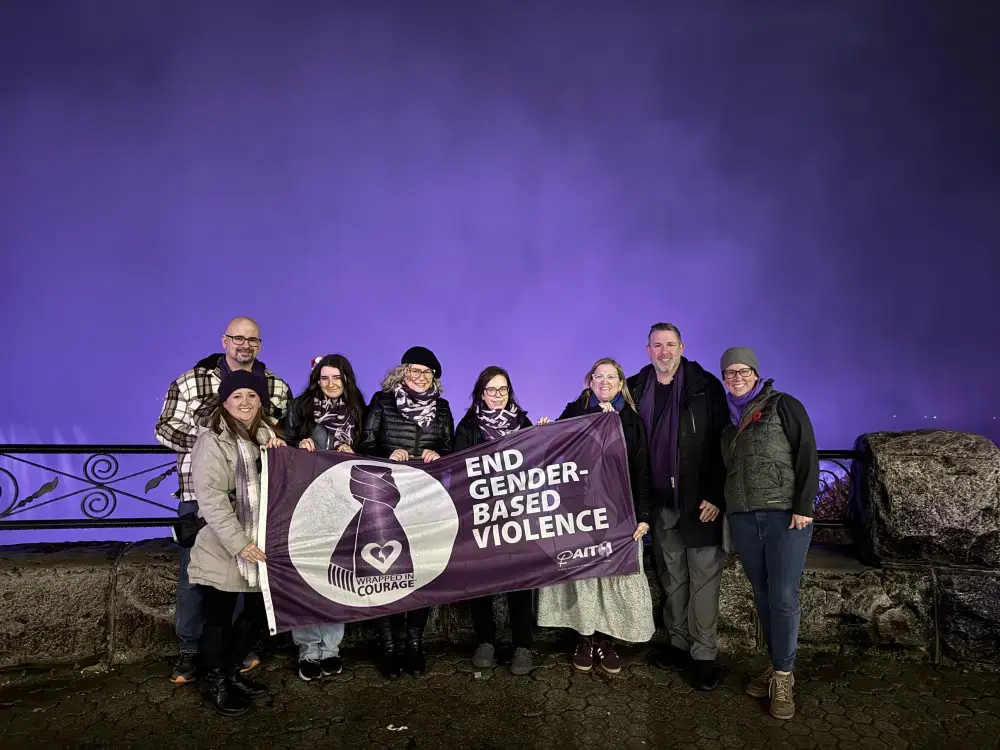 A group of people stand in front of Niagara Falls, which is glowing purple, with a flag that says End Gender-Based Violence.